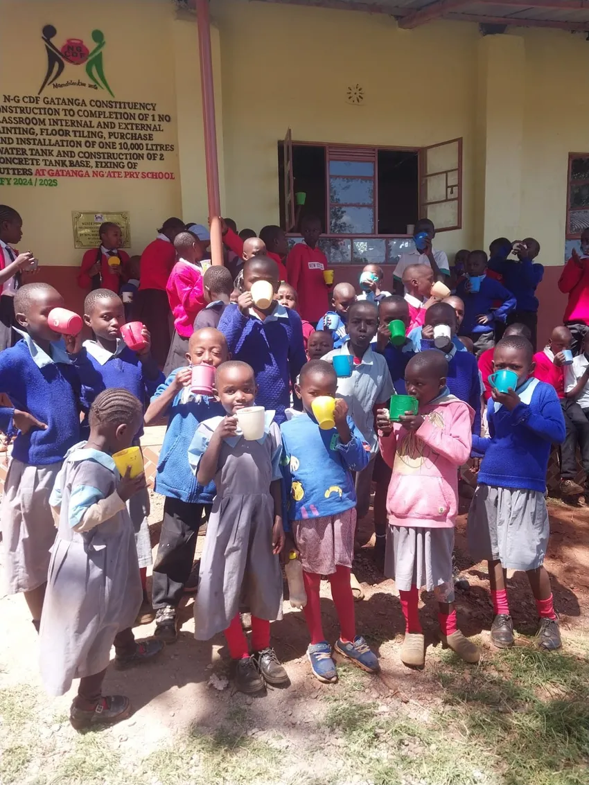 A large group of approximately 30 Kenyan children holding plastic mugs of porridge, standing outside a Kenyan school.