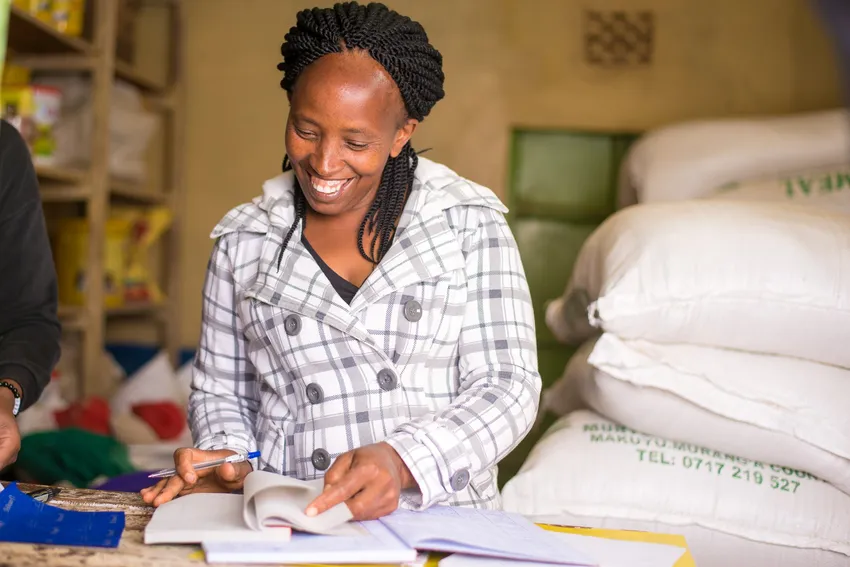 A woman records information in a notebook beside sacks of goods in her shop.
