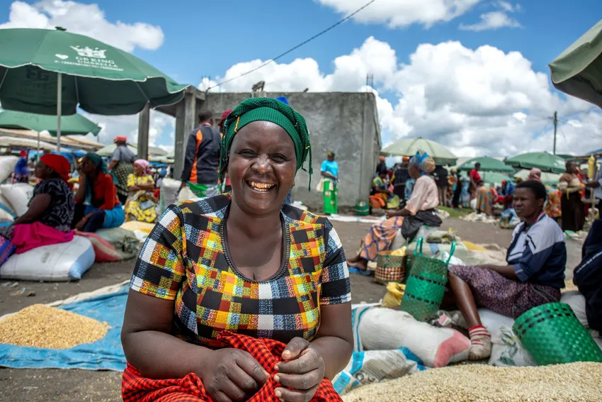 A woman sits smiling at a busy market, with bags of grain around her.