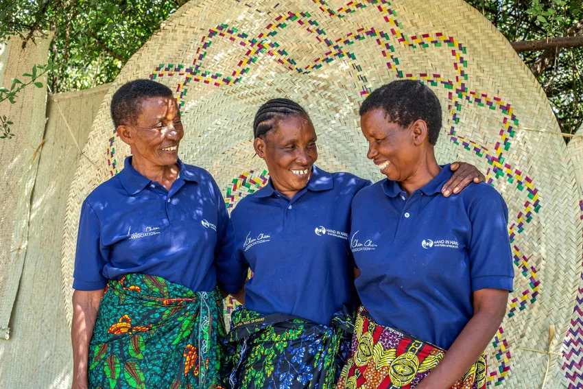 Three women smile together beside their handmade woven baskets and mats.