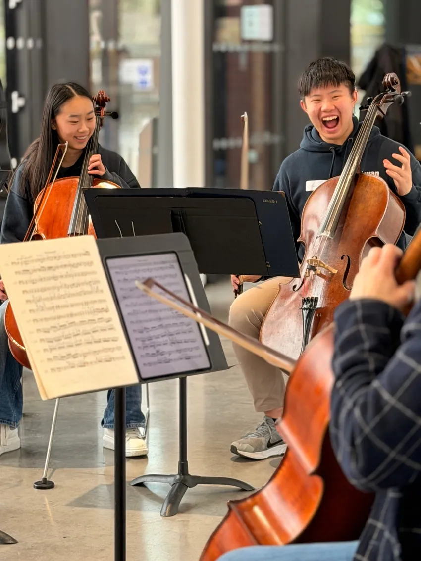 Two young musicians are sat down playing the cello, one smiling and the other is laughing.