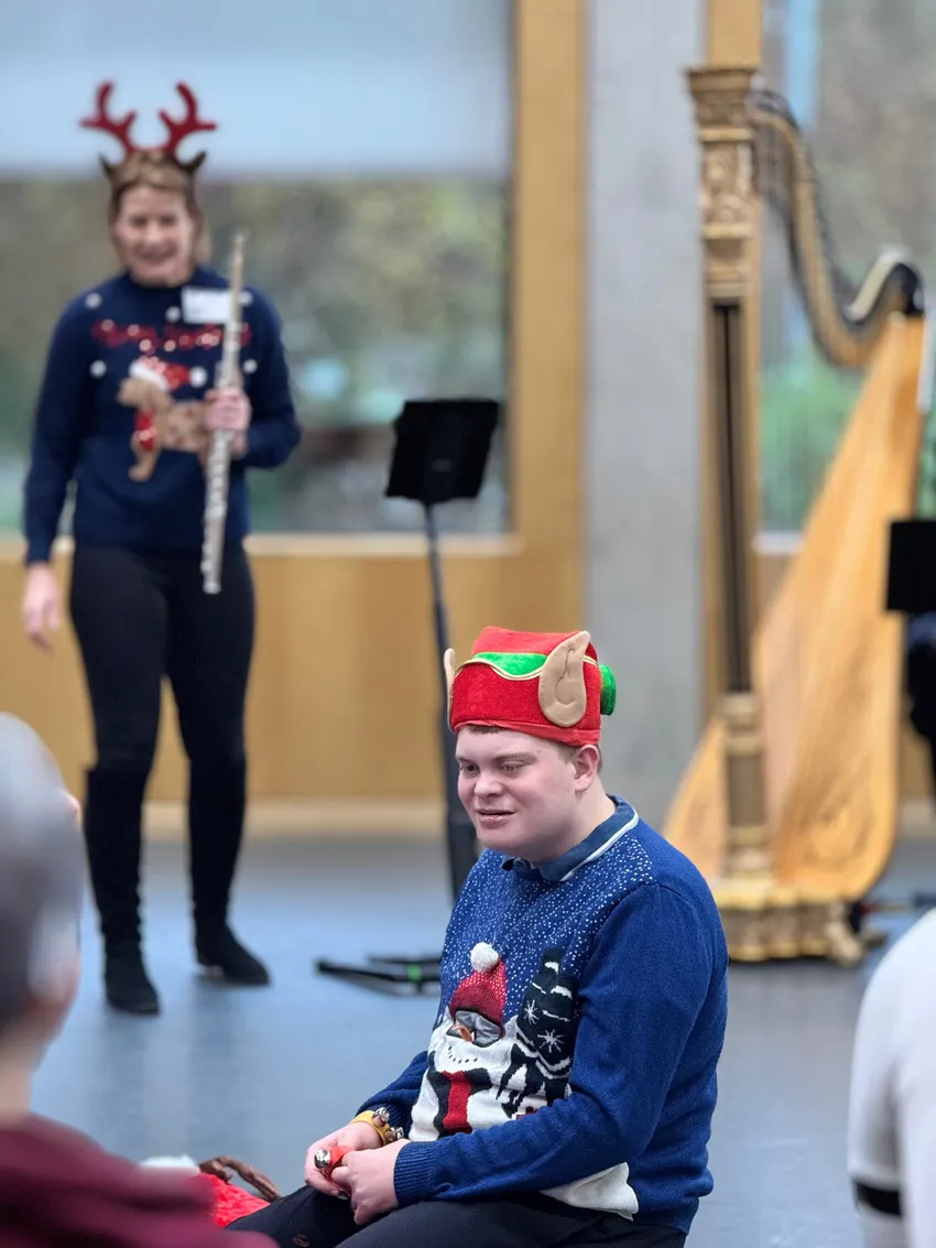 Young person wearing a Christmas hat with elf ears is kneeling in front of a musician holding a flute wearing antlers and a harp. Everyone is smiling.