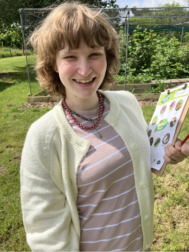 A woman is smiling wearing is wearing a cardigan over a striped top and necklace. She is holding a clipboard with a nature-themed activity sheet at Flower Pod Newark