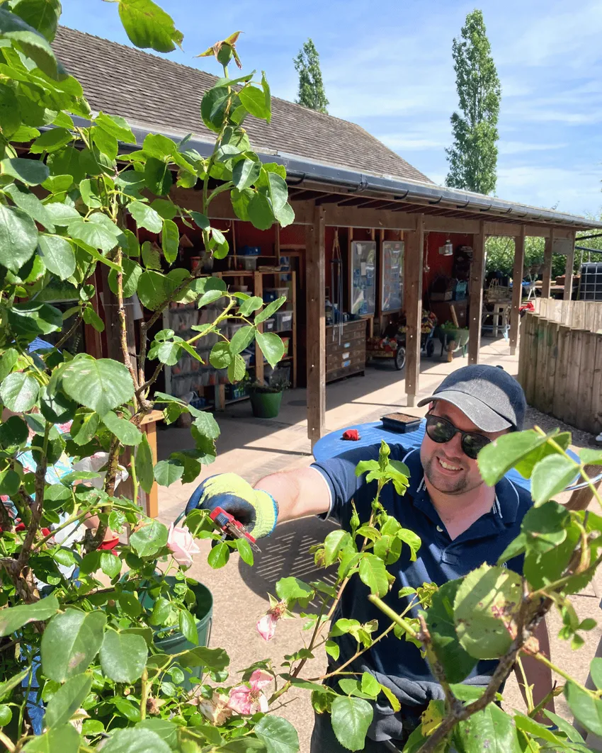 Man smiling wearing cap, sunglasses and gloves while deadheading a rose bush outside Flower Pod Southwell building.
