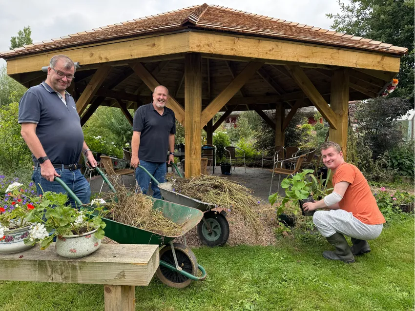 3 men smiling while gardening in front of a wooden gazebo, using wheelbarrows and planting flowers at Flower Pod Southwell