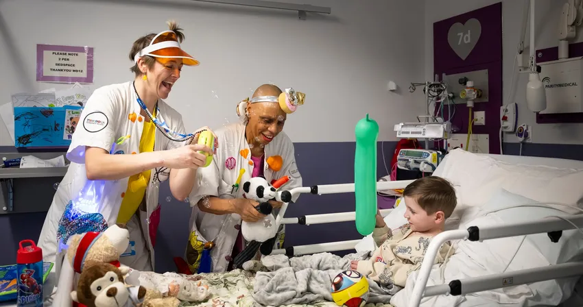 Dr Dilly Dally & Dr Geehee are playing with a child in a hospital bed. Dr Dilly Dally is using a bubble machine, Dr Geehee is holding a teddy panda and the child is holding a green balloon.