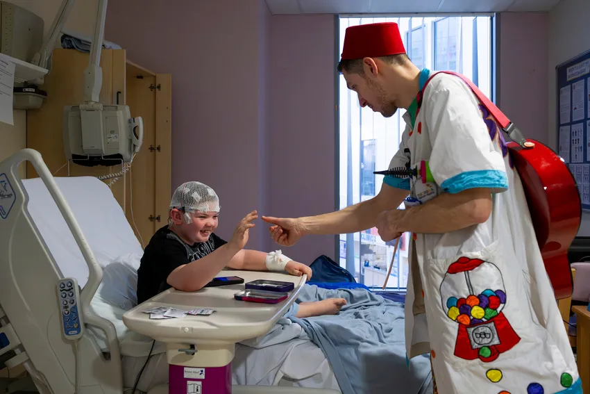 Dr Bungee is interacting by an older child who is sitting in a hospital bed. The child is holding his hand up and Dr Bungee is reaching his thumb out