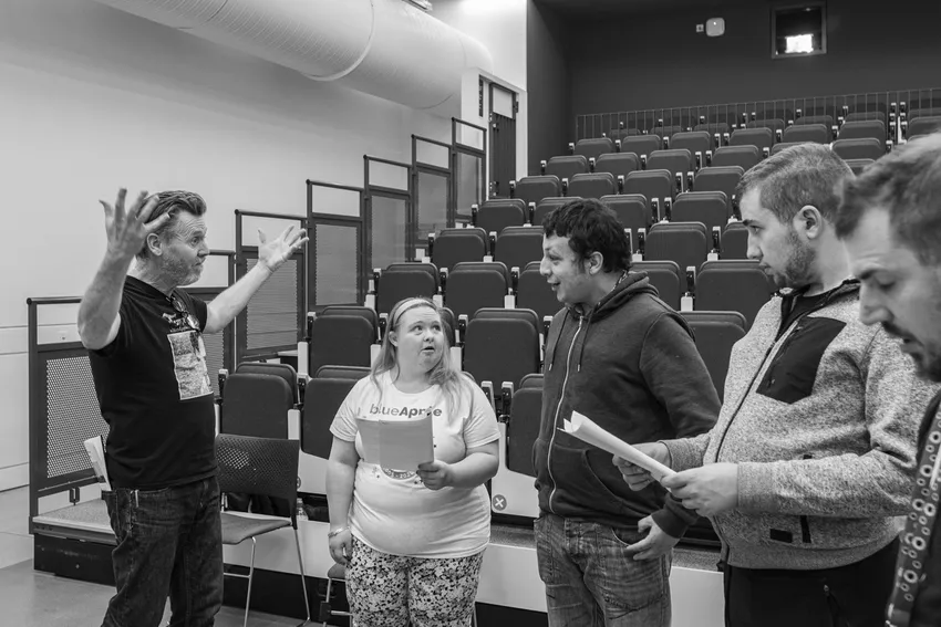 Black‑and‑white photo of a small group standing in a lecture theatre. One person at the front is speaking with raised arms. Three others are standing nearby holding papers, and rows of empty tiered seats fill the background.