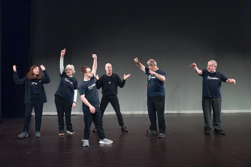 A group of people on a stage, each with one arm raised or gesturing as they rehearse together against a plain backdrop