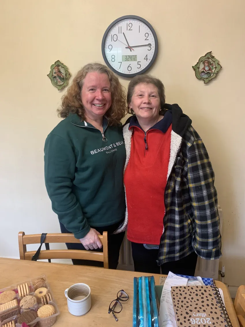 Two ladies standing behind a kitchen table with a cuppa and biscuits on the table. They are both smiling at the camera.