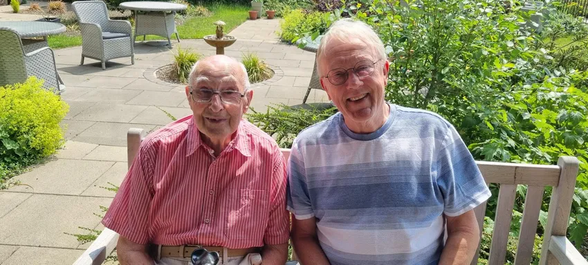 Two gentlemen are sitting on a bench in a garden in the sunshine - they are both smiling at the camera