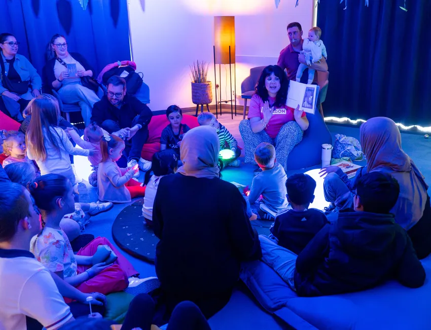 A group of families sat listening to a story, under blue lights