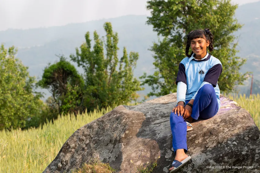A young girl in school uniform sitting on a rock in a field in India, smiling confidently at the camera.