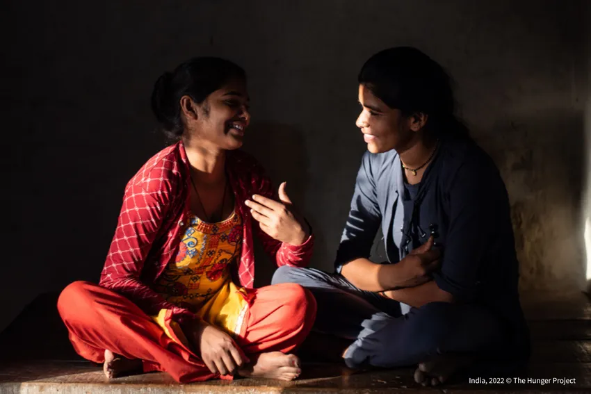 Two adolescent girls sitting together in India, engaged in conversation, one gesturing expressively while the other listens.