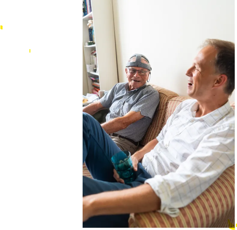 On the left, John wears the Fastball cap as he takes part in at-home trials for the BRACE-funded early diagnostic tool. He is laughing with Dr George Stothart, who is on the right and invented the test, which is being trialled by the NHS.