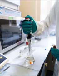 A gloved hand in a science lab lowers a pipet into a glass container containing a clear liquid chemical