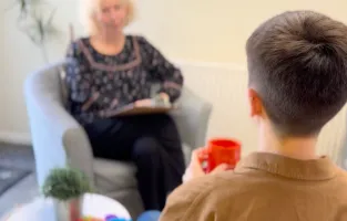 A young person holding a cup of tea receives one-to-one holistic support from a Centre 33 staff member, seated together in a calm, welcoming space.