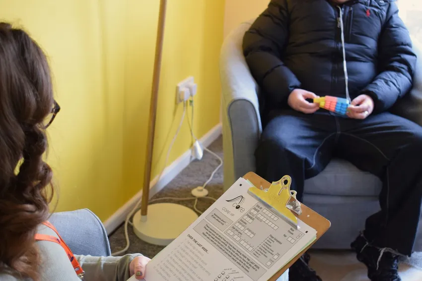 A young person speaks with a Centre 33 Engagement and Support Project Worker during a drop-in triage session. The worker holds an assessment form while the young person sits opposite, holding a puzzle toy for sensory support
