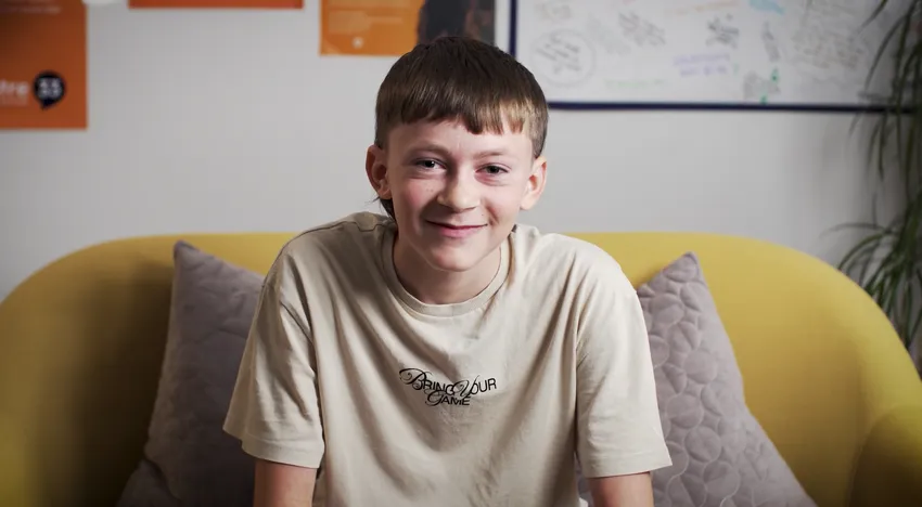 Arthur, a young carer, smiles at the camera while seated on a yellow sofa in a welcoming Centre 33 space.