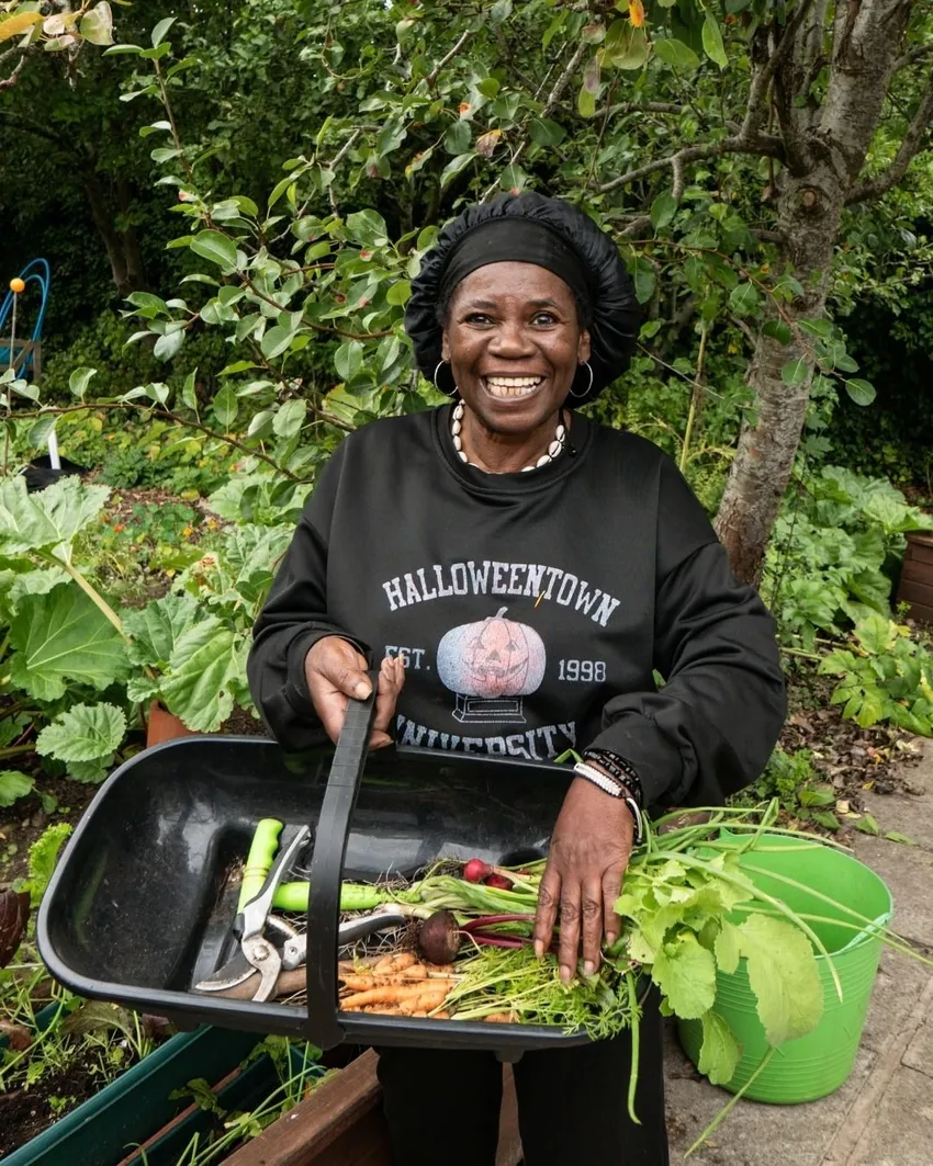 Client gardener, Ciceley, proudly holds a tray of freshly harvested vegetables in the garden.