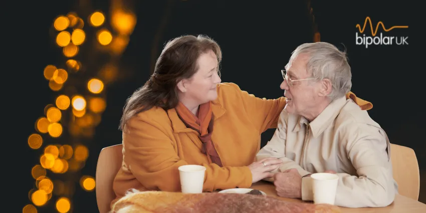 Warm orange lights in the background with an elderly man sitting at a table looking at a woman who is helping him.