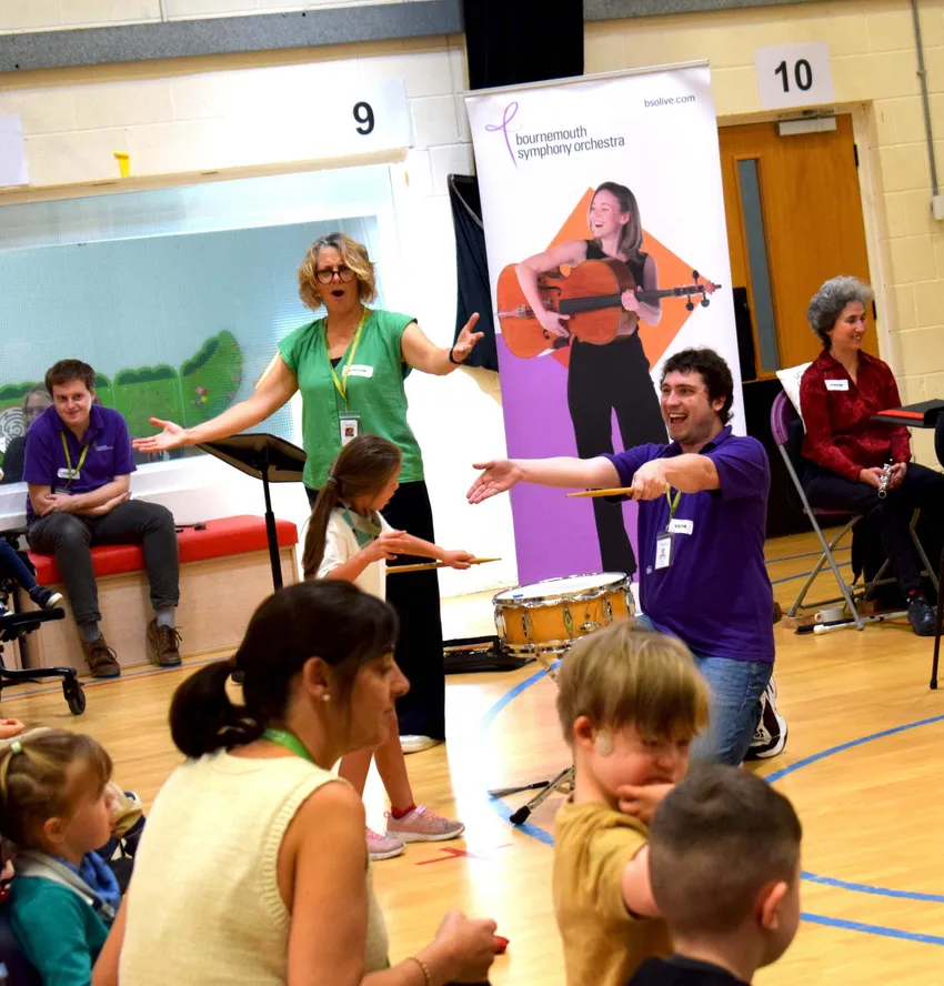 In a school hall, two BSO musicians - one man, one woman - are in front of an audience of children, holding their arms out in front of them as they sing. The man kneeling on the floor has a drum in front of him.