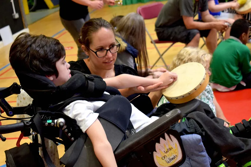 A young boy in a wheelchair holds a tambourine on his lap, while a teacher kneels beside him to assist in tapping it.