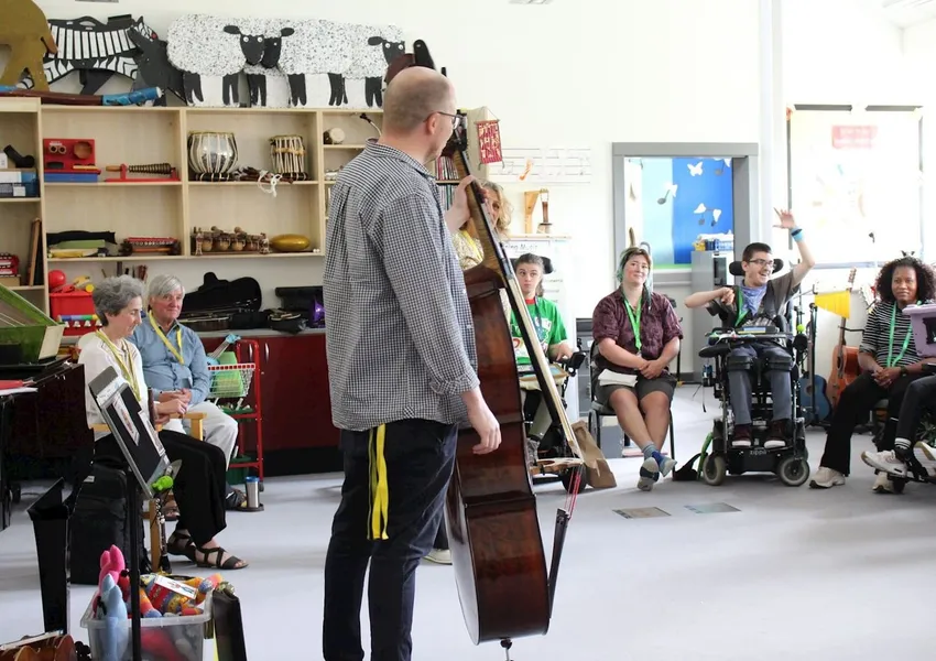 A man who is a BSO musician stands in the centre of a circle of students, holding his double bass which is the same height as him. In the background, a student in a wheelchair watching on is smiling and raising his hands.