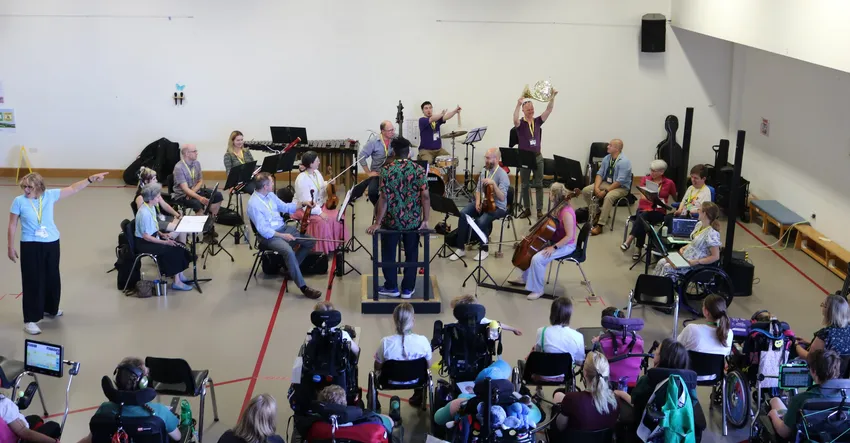 In the foreground students of the SEND school sit and watch the 14-piece Orchestra as a musician stands up to present his French horn.