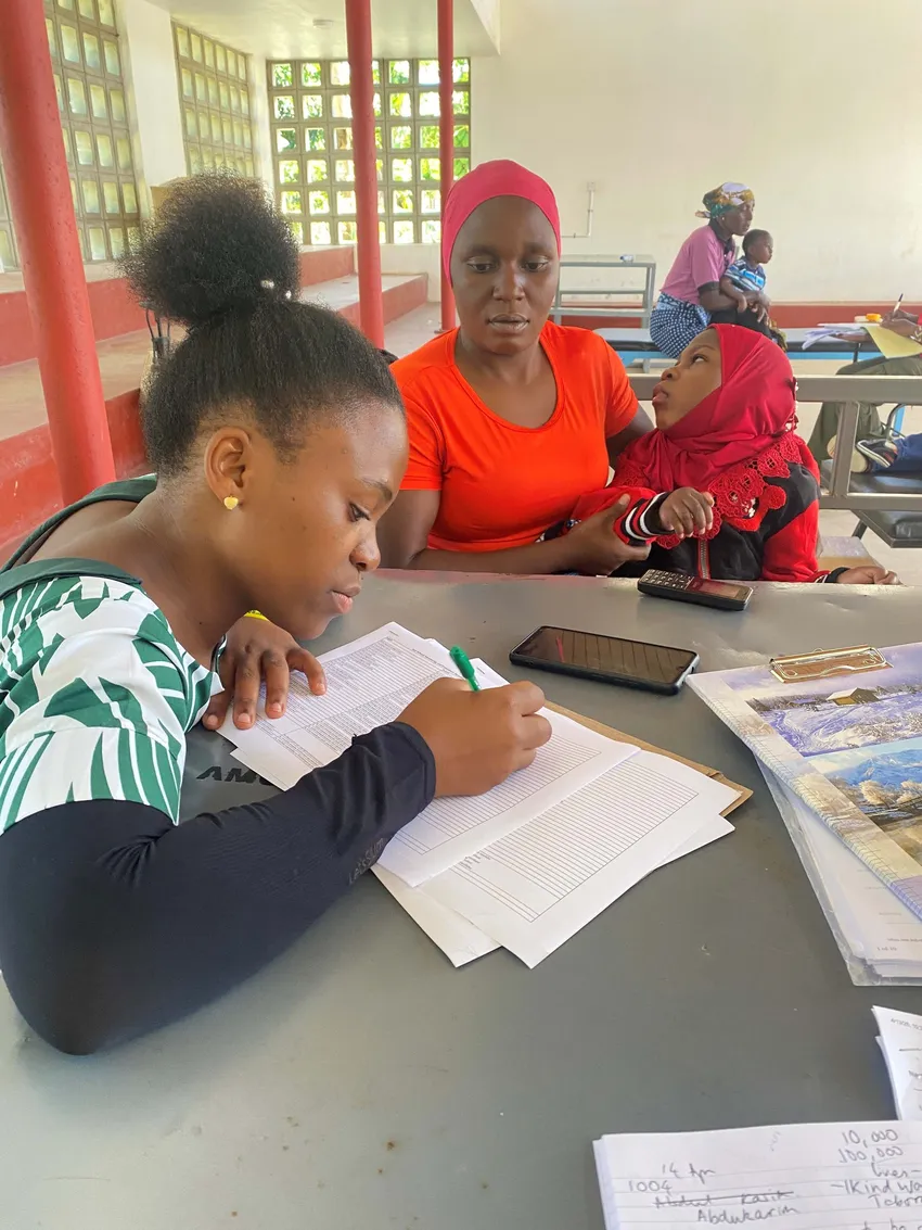 A young black woman is filling out a monitoring and evaluation form. Beside her a mother and daughter sitting on her knee