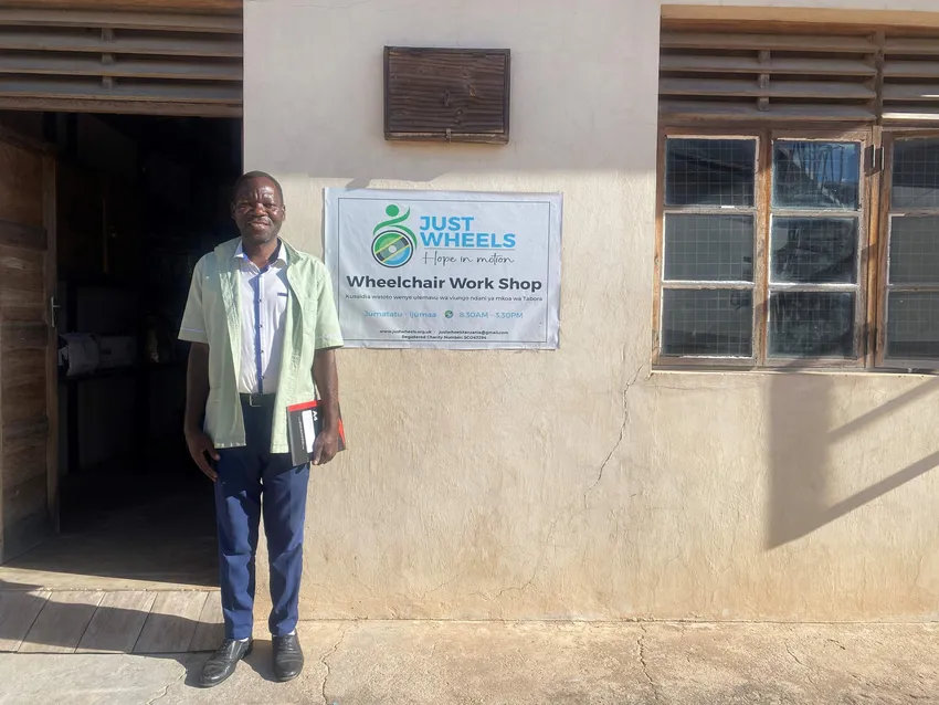 Edgar, our wonderful wheelchair technician outside his new workshop. ALL black male stands beside a sign and an open door. the sign says just wheels wheelchair workshop