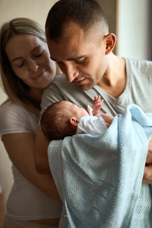 Dad holding baby. Mum in background smiling over shoulder