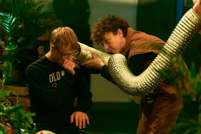 An actor holds a long silver tube to a young person's ear.