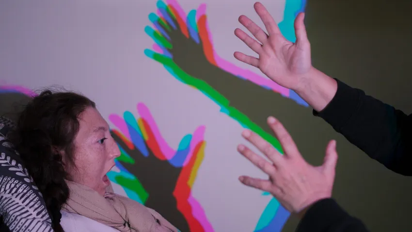 Young person looks at another person's hands. Behind there are multi-coloured shadows.
