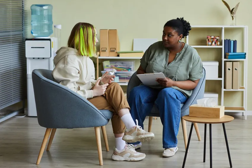 Image 1 (iStock)  "A young woman with green highlighted hair sits in a grey armchair talking to a professional who is holding a clipboard, in a calm office setting
