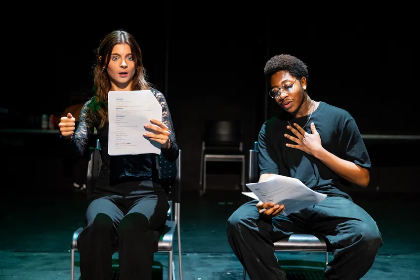 Two students in black sitting on chairs reading from a script