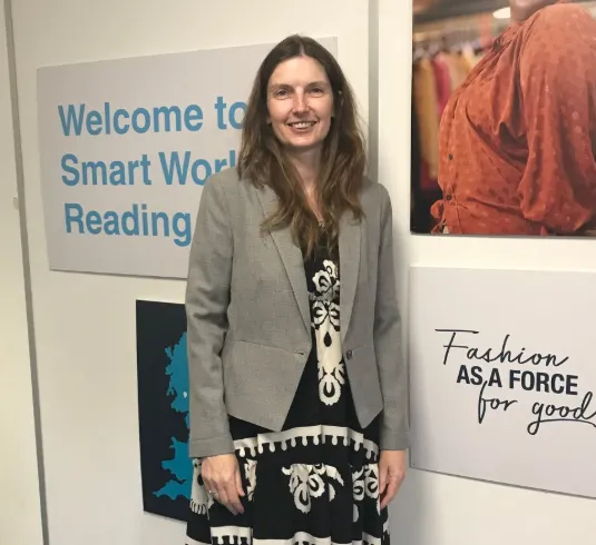 Smart Works Reading Client Emma stands next to the Smart Works Reading sign in her interview outfit provided by Smart Works Reading. Next to her, a sign reads "fashion as a force for good".