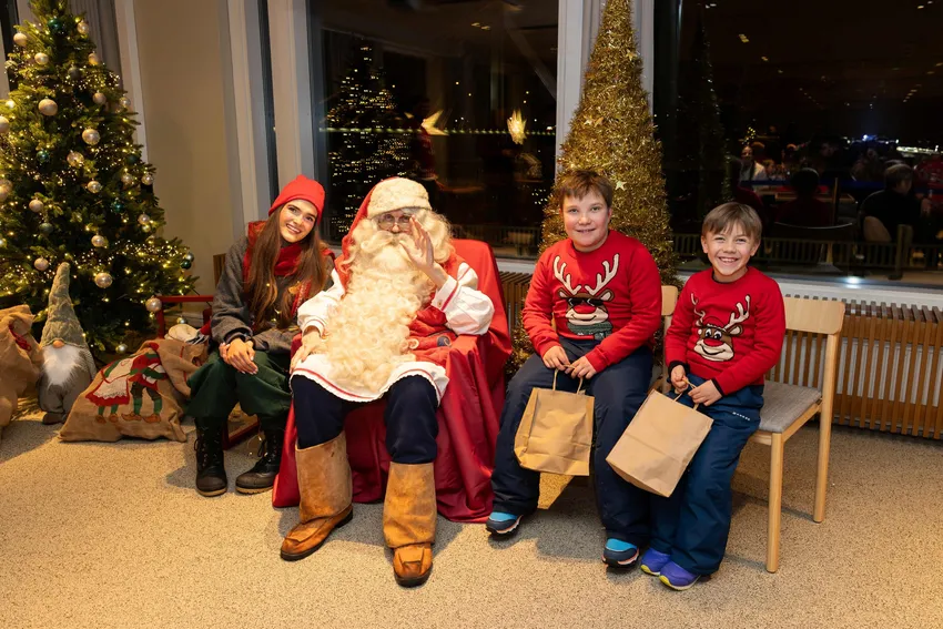 Two children in matching reindeer sweaters sit smiling beside Santa and an elf in a warmly lit Christmas room.