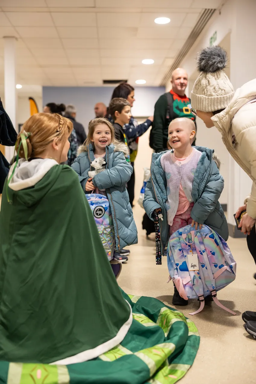 Two excited children with rainbow backpacks talk to a cloaked volunteer in a busy indoor queue, smiling as they wait to start their Lapland trip.