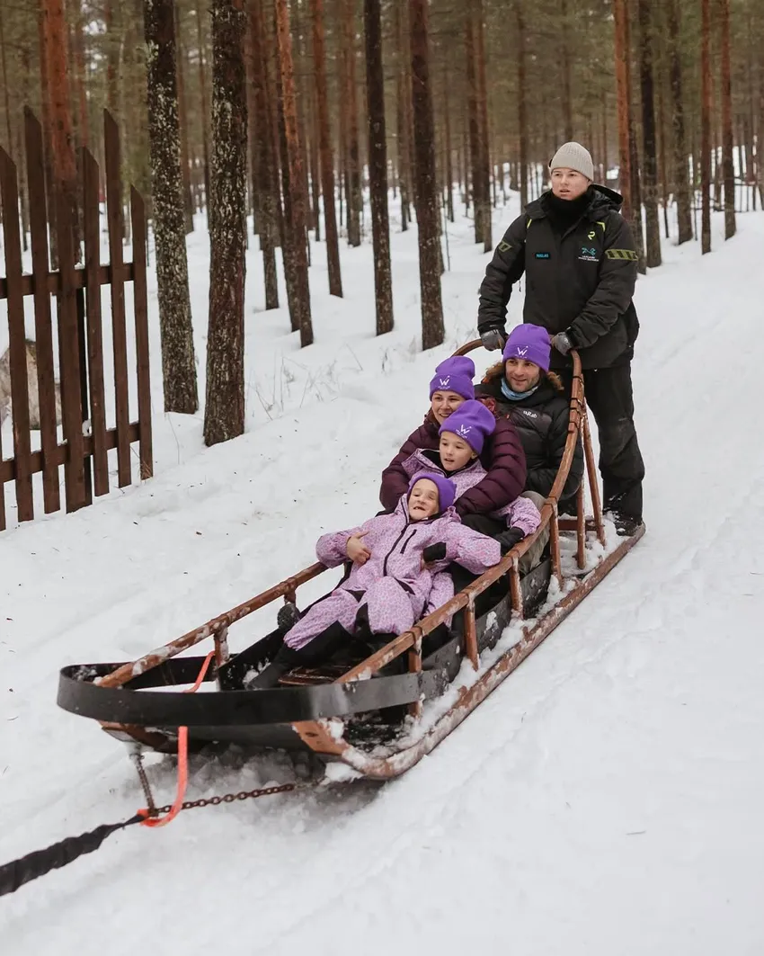 A guide pulls a long wooden sled through a snowy forest, carrying a bundled‑up family in matching purple winter clothes and hats.