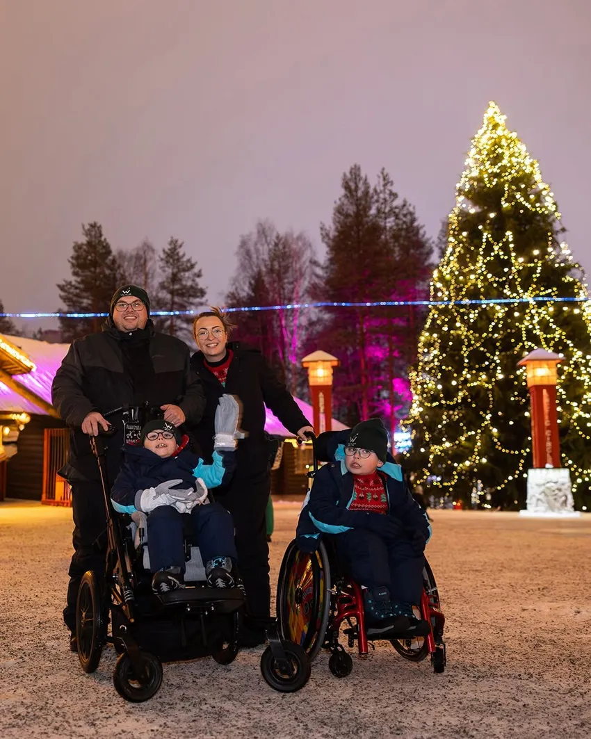 Two parents pose smiling with two children in wheelchairs in front of a towering Christmas tree lit with golden fairy lights in a snowy Lapland village.
