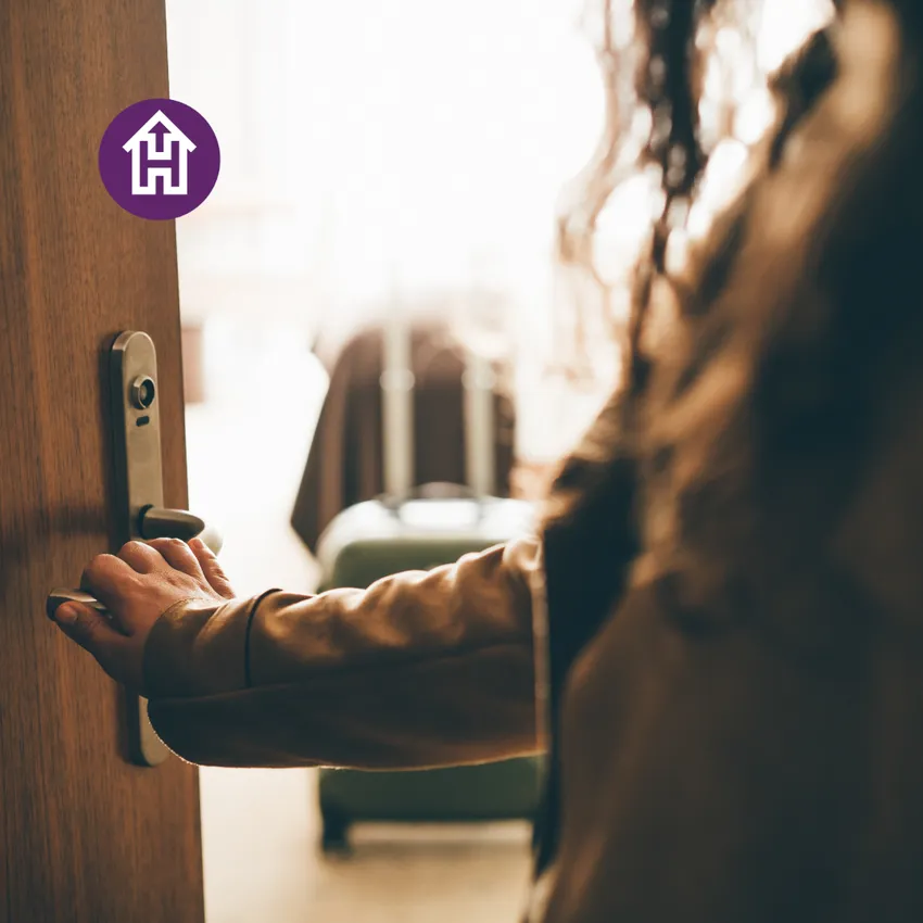 A person opening a door to a room showing a bed and suitcase, symbolising safe accommodation and a fresh start