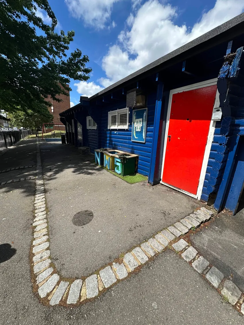 NW5 Hut community building in Kentish Town, a blue wooden building with a red door and white-framed windows beside a paved path, trees and a playground.