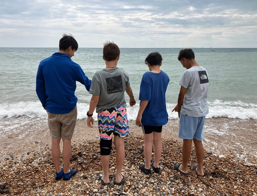 Four young people stand on a rocky beach looking out at the choppy, blue green ocean.