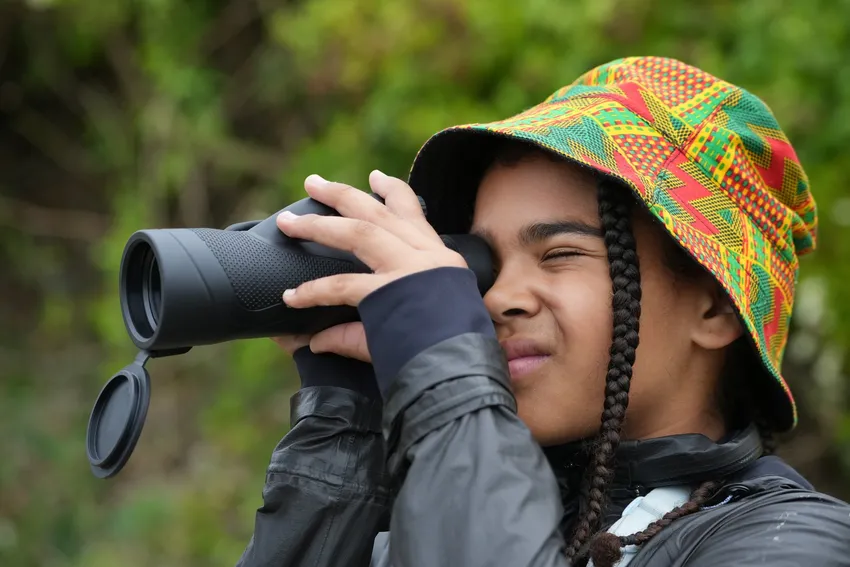 A young person wears a colourful patterned bucket hat and looks through a monocular, the background is a green outdoor space.