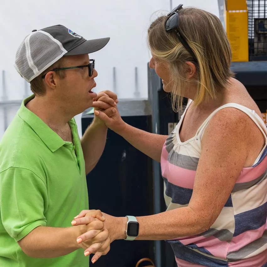 A young, white man in a green shirt and a white and grey baseball cap is facing a white lady with blond hair and sunglasses on her head. They are holding hands, both hands, with fingers interlinked and they are gazing directly at each other.