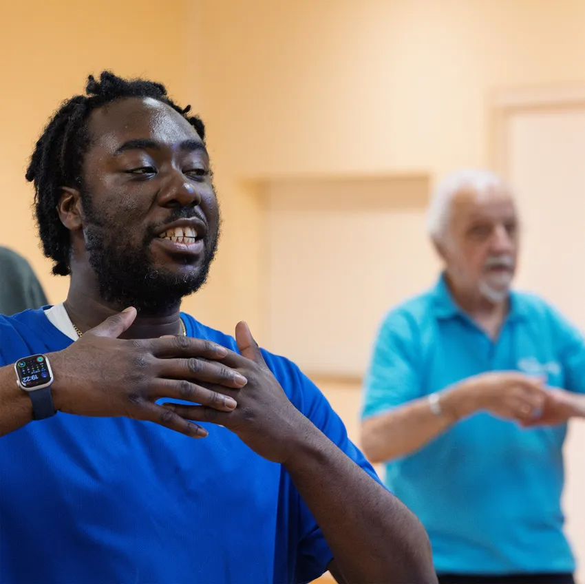 A young, smiling black man in a blue shirt is gesturing with his hands. Behind him, an older white man is visible doing the same gesture.
