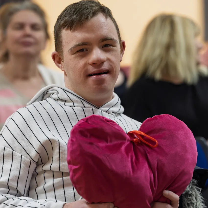 Young man in white, striped hoody, holding a red velvet heart. He is smiling at the camera. He has short dark hair; he has Down Syndrome. There are 2 ladies visible, though blurred, in the background, also smiling