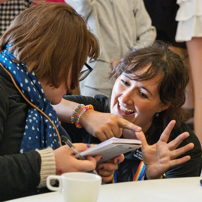 A young lady with brown hair and glasses is showing another lady with dark hair her notebook. The second lady is smiling broadly and signing, as though she understands and is pleased.