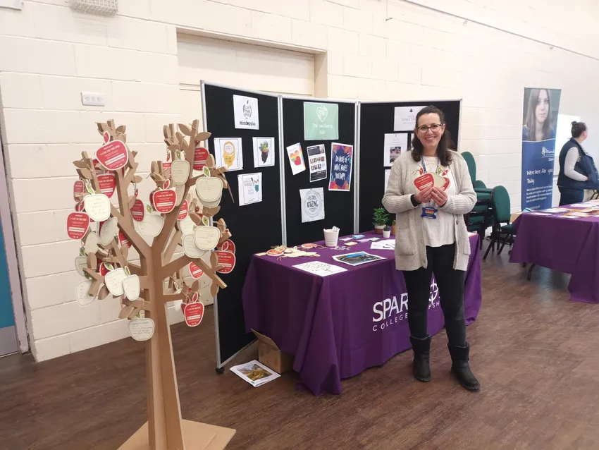 College tutor stands next to a wooden mindapples tree, holding red and cream apple shaped cards at a student welcome week